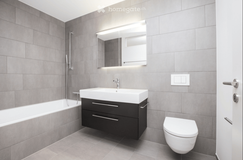 Modern bathroom with grey tiled walls and floor, featuring a bathtub, sink with a black cabinet, toilet, and a mirrored cabinet with built-in lighting above the sink.