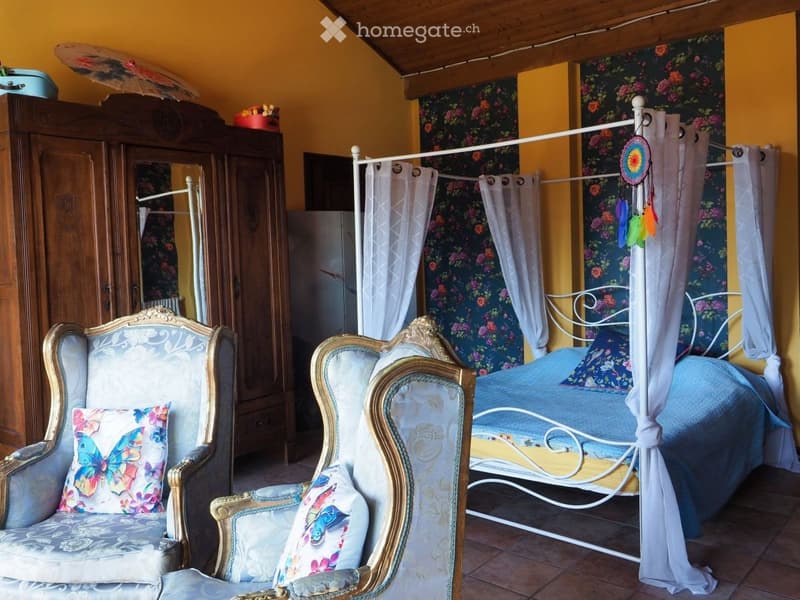 Cozy bedroom with a four-poster bed, two armchairs, a wardrobe, and a mirror. The room features floral wallpaper, colorful pillows, and a dreamcatcher hanging from the bed frame.