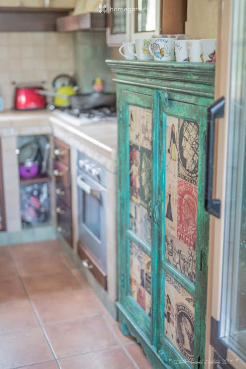 A kitchen with a green, vintage-style cabinet adorned with colorful posters, a stove with a teapot, and several cups on the counter.