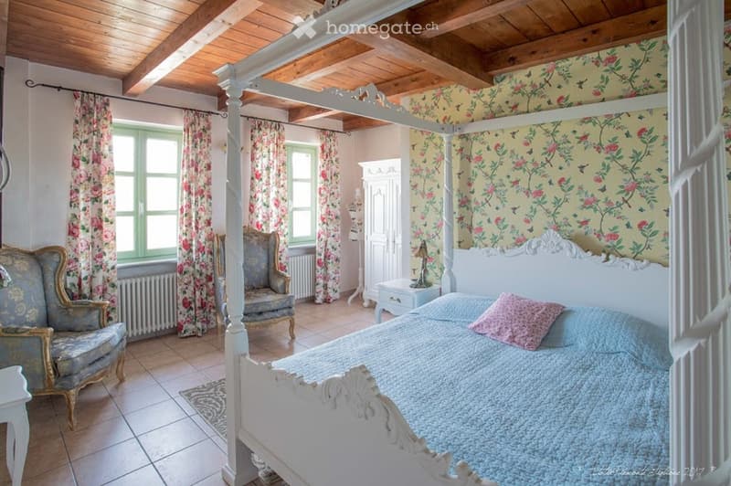 Elegant bedroom with a four-poster bed, floral wallpaper, two armchairs, nightstand with lamp, and radiator.