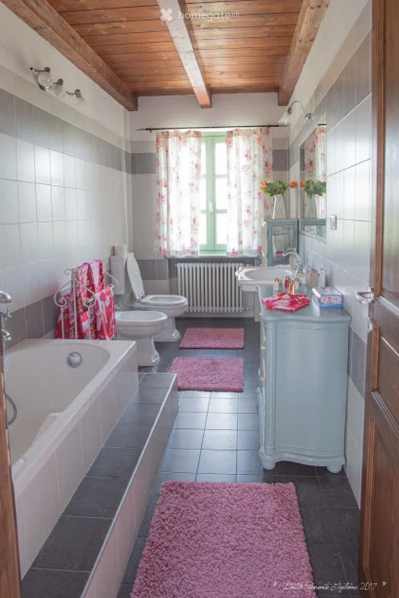 Bathroom with a bathtub, two toilets, a sink, a vanity, a radiator, a mirror, towels, a curtain, and a vase with flowers.