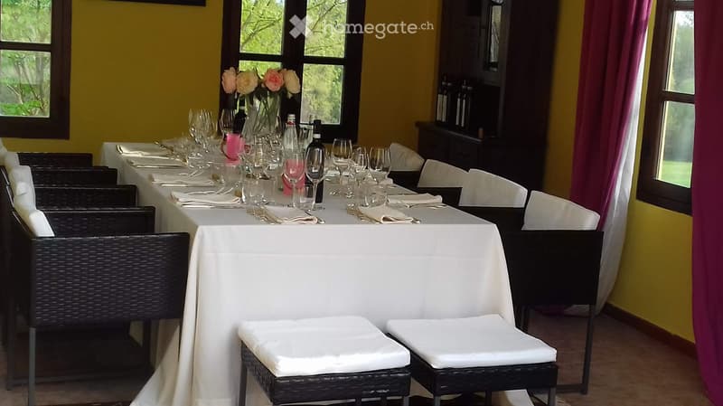 A long dining table set with white tablecloths, wine glasses, silverware, and napkins, surrounded by black wicker chairs with white cushions. A vase with pink and white flowers is placed at the center of the table.