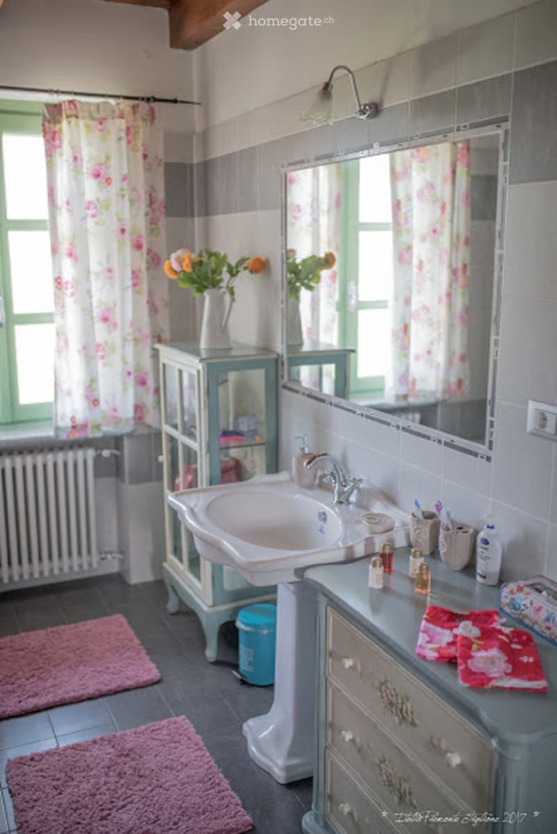 A vintage-style bathroom with a white sink, mirror, floral curtains, a small cabinet with a vase of flowers, and a vanity with toiletries and towels.