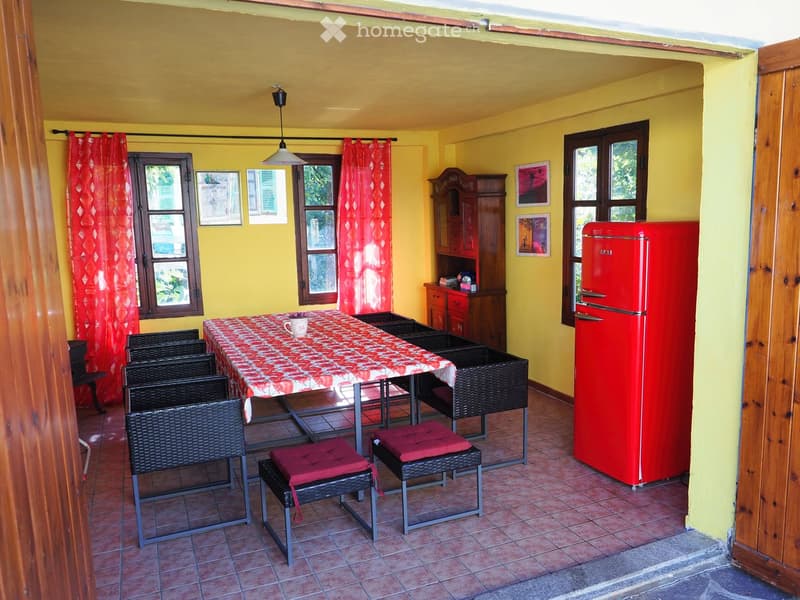 Bright kitchen with yellow walls, red refrigerator, dining table with floral tablecloth, wicker chairs, and wooden stools.