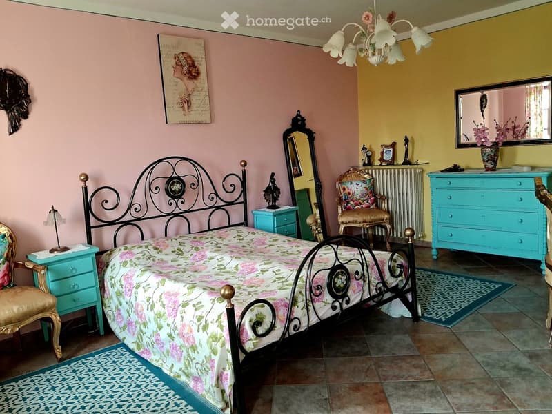 A well-decorated bedroom with a floral bedspread, ornate metal bed frame, two nightstands, a floral armchair, a dresser, a mirror, and decorative items.