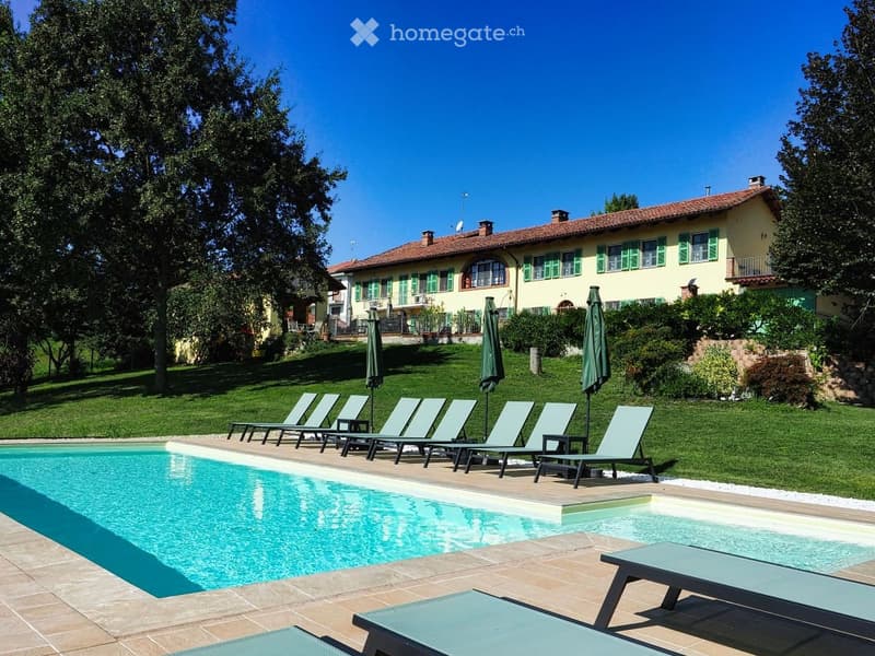Outdoor area with a swimming pool, lounge chairs, umbrellas, and a large house with green shutters in the background.