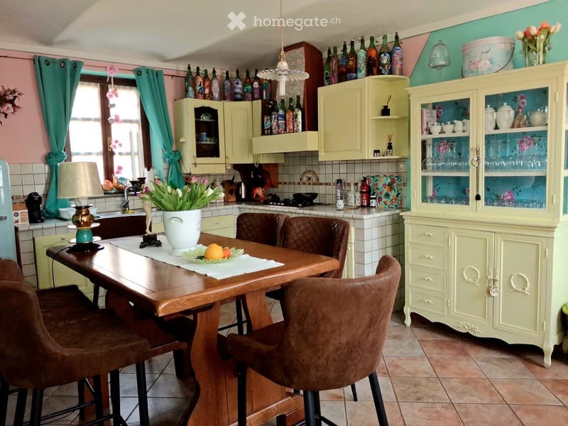 Bright kitchen with pastel-colored cabinets, a wooden dining table with four brown leather chairs, a vase with flowers, and various decorative items including bottles and glassware on the cabinets.