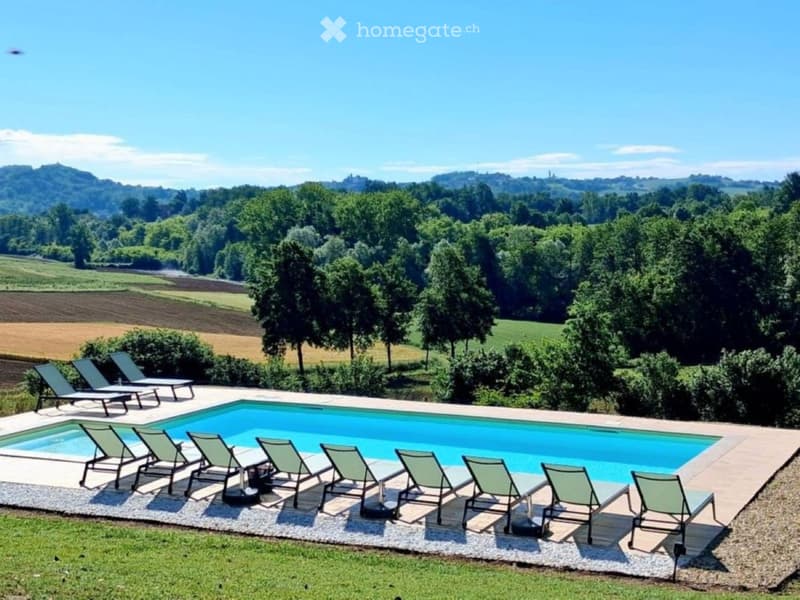 Outdoor area with a swimming pool surrounded by lounge chairs, set against a scenic countryside backdrop with trees and fields.