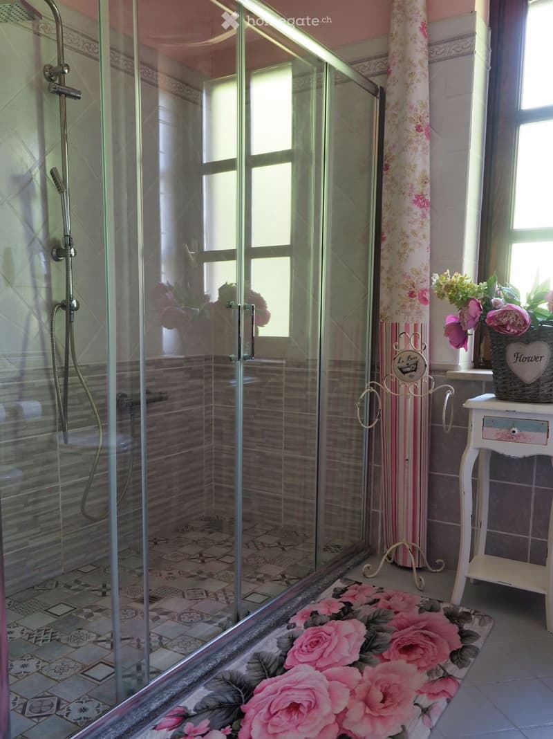 Modern bathroom with a glass-enclosed shower, decorative floral rug, small white table with flower arrangement, and floral curtain.