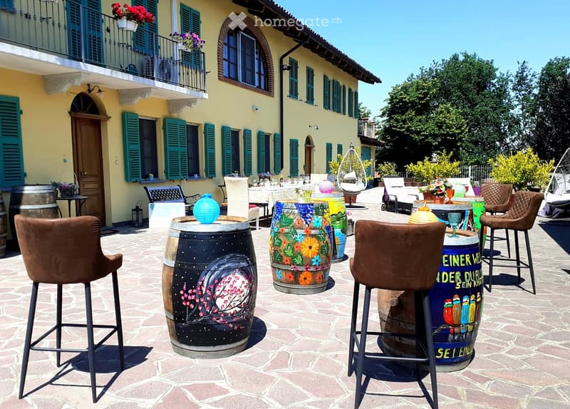 Outdoor patio area with stone flooring, several chairs and tables, decorative barrels used as tables, and a balcony with shutters and potted plants.