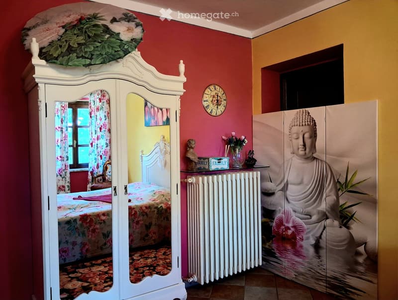 A vibrant bedroom with a floral-patterned bed, a large white wardrobe with mirrors, a radiator, a decorative clock, and a screen displaying a Buddha statue.
