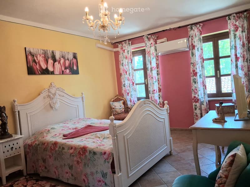 A well-furnished bedroom with a large bed, nightstand, chair, table, and floral curtains. The room features a chandelier and a picture frame on the wall.