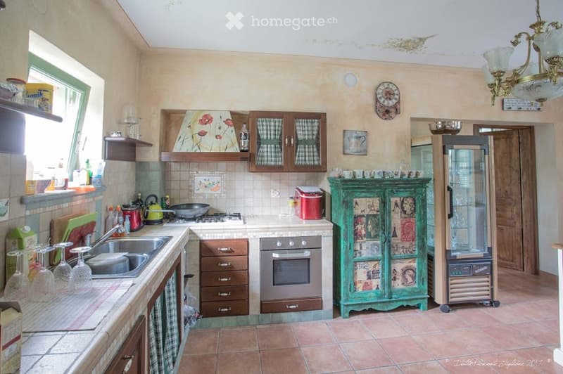 The kitchen features wooden cabinets with checkered curtains, a stainless steel oven, a sink with a faucet, and various kitchen items including wine glasses, a teapot, and a frying pan. The floor is tiled, and there is a decorative green cabinet and a ref