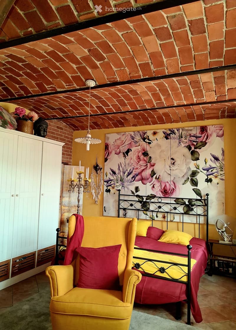 A cozy bedroom with exposed brick walls and a brick ceiling. Features a metal-framed bed with red and yellow bedding, a yellow armchair, a white cabinet, and a decorative candle holder.