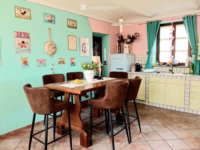 Brightly colored kitchen with a wooden dining table surrounded by brown leather chairs, a vintage-style refrigerator, and a sink area with a window. The walls are painted in pastel green and pink, decorated with framed pictures and a clock.
