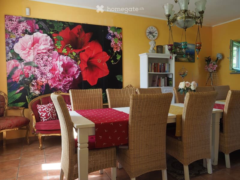 Brightly colored room with a floral screen, a red sofa, a dining table with wicker chairs, a bookshelf, and decorative items including a chandelier and a clock.