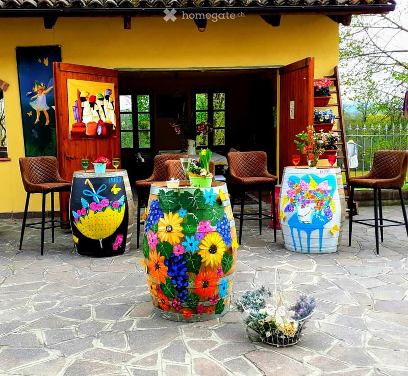 An outdoor patio area with stone flooring, colorful painted barrels used as tables, chairs, and flower pots.