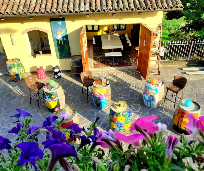 Courtyard with stone pavement, colorful painted wine barrels used as tables and chairs, small flower arrangements, and a view of the interior dining area.