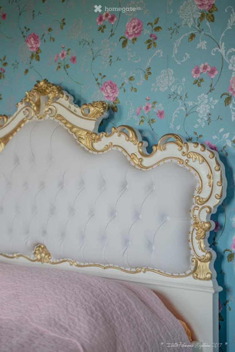 Elegant bedroom with a white and gold ornate headboard, tufted white upholstery, and floral wallpaper in shades of blue and pink.