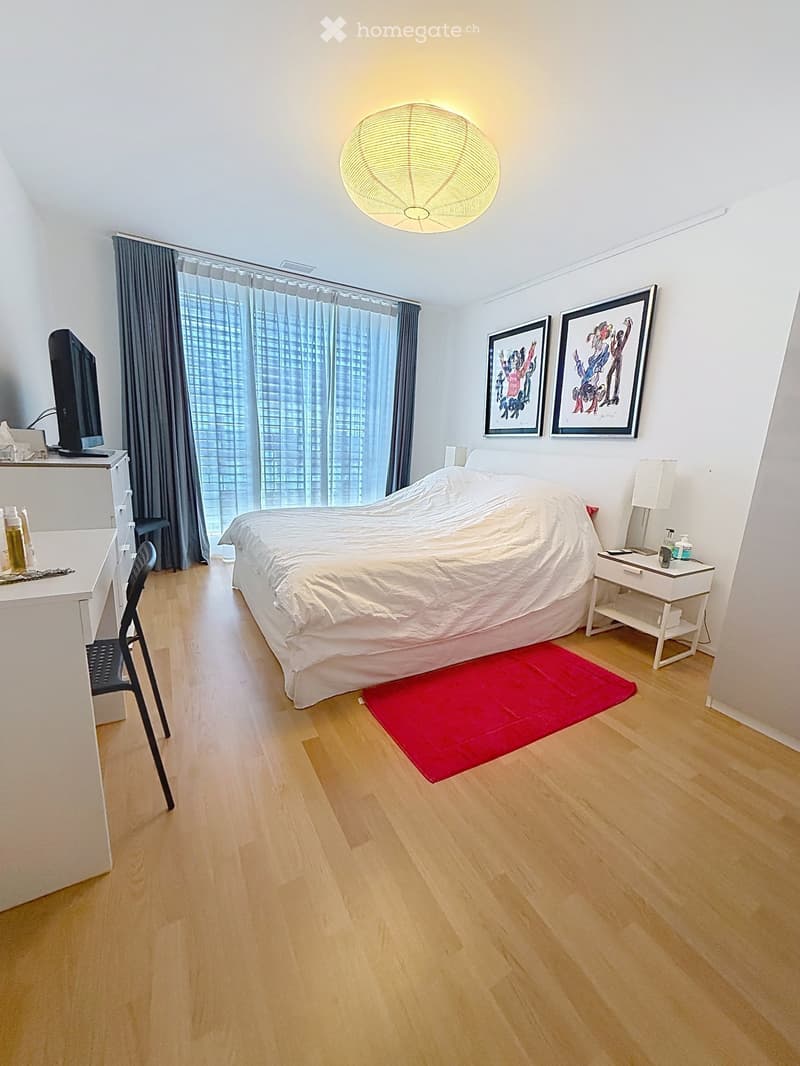 A modern bedroom with a large bed, a red rug, a chair, a small table with a lamp, a screen, and two framed pictures on the wall. The room has wooden flooring and large windows with curtains.