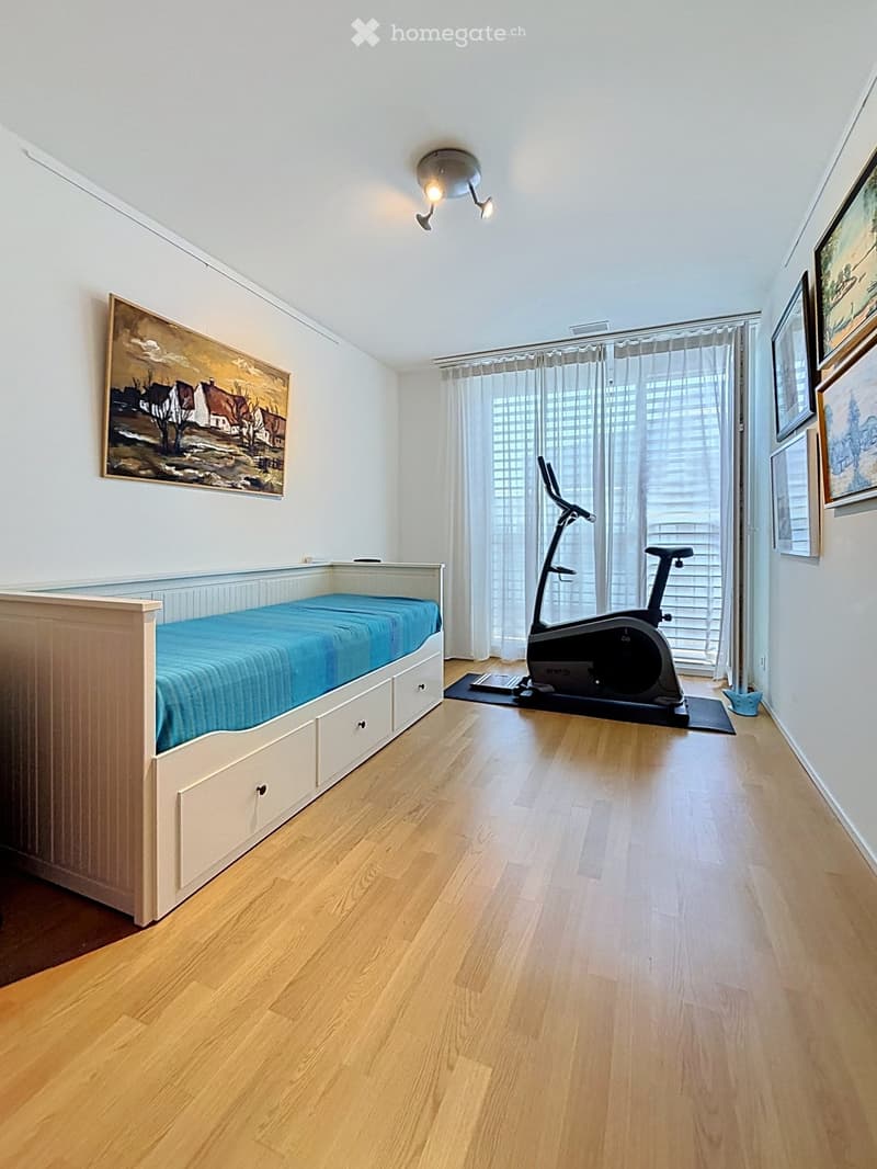 Well-lit bedroom with wooden flooring, a built-in bed with drawers, an exercise bike, and framed artwork on the walls.
