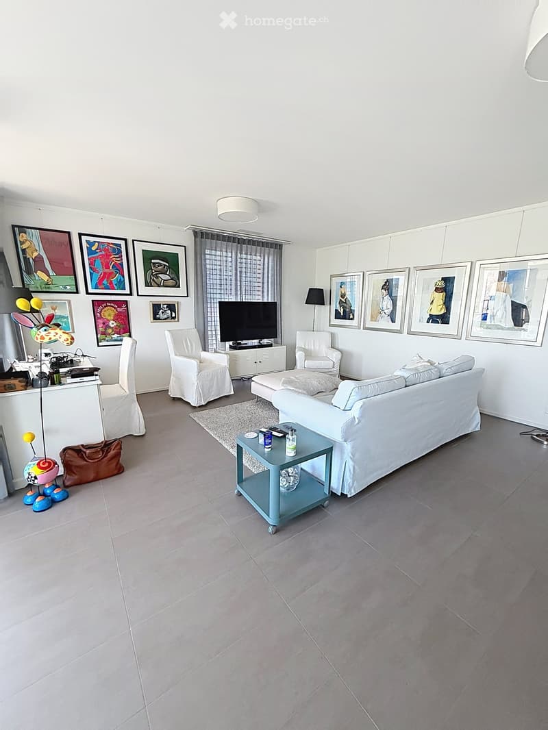 Bright living room with white walls, grey tiled floor, large sofa, coffee table, armchairs, ottoman, TV, and decorative artwork.