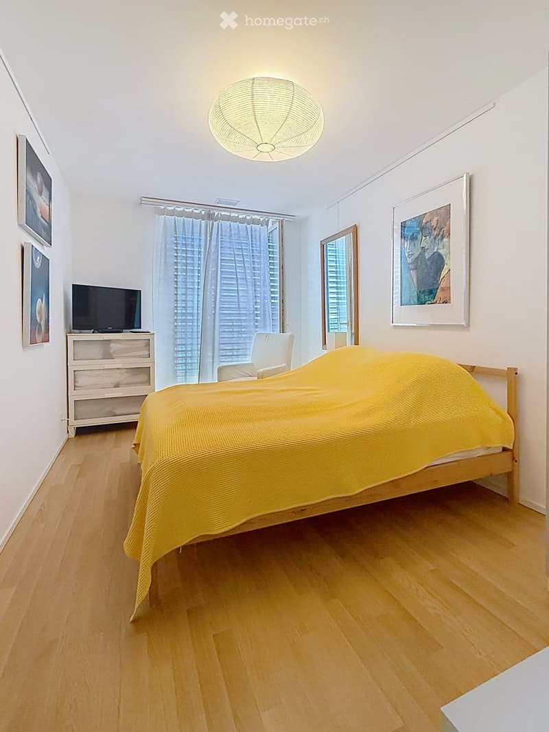 Bright bedroom with wooden flooring, a large bed with a yellow blanket, a white chair, a television on a cabinet, and several picture frames on the walls.