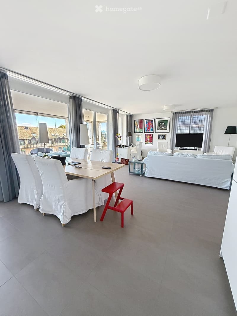 Open-plan living and dining area with large windows offering a view, featuring a white sofa, dining table with chairs, a red stool, and a television.