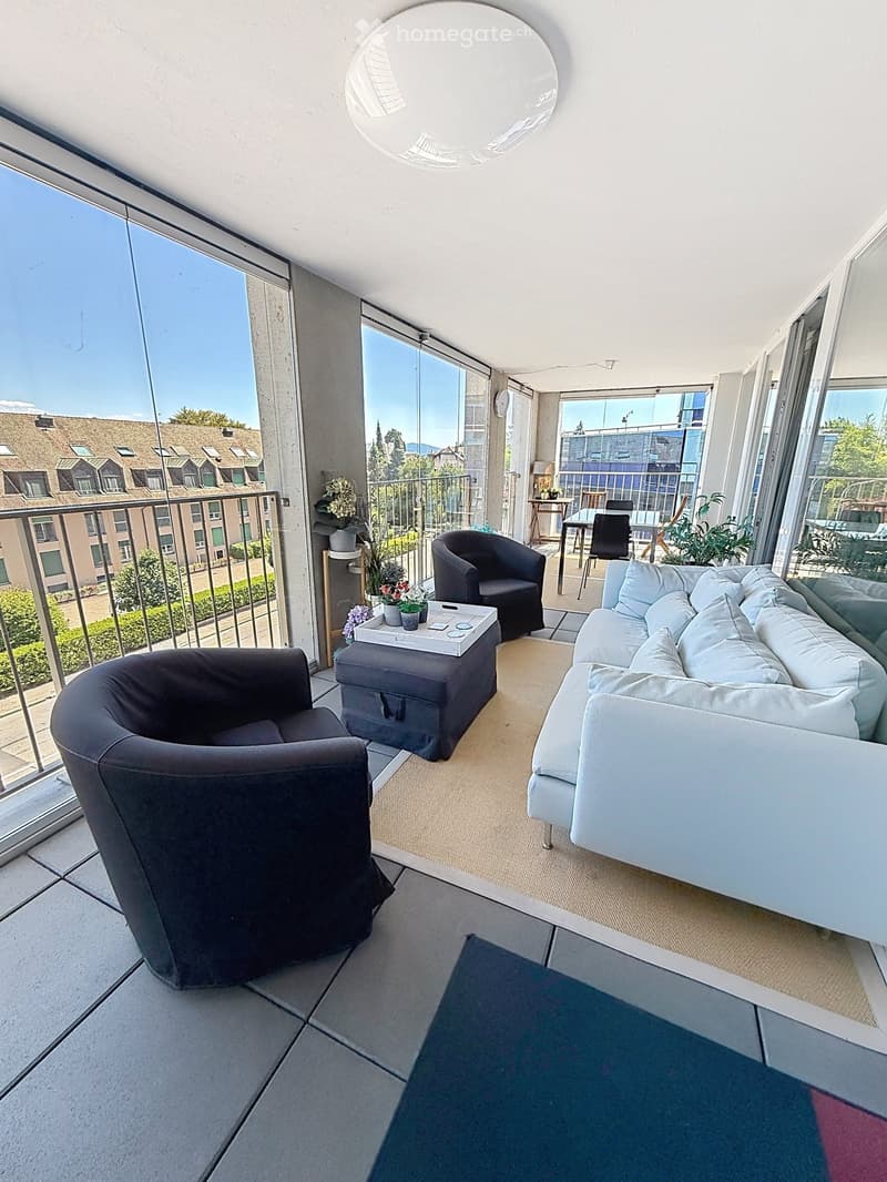 A modern, furnished balcony with a white sofa, two black armchairs, a small table, and a grey ottoman. The space features large glass windows and a view of the city.