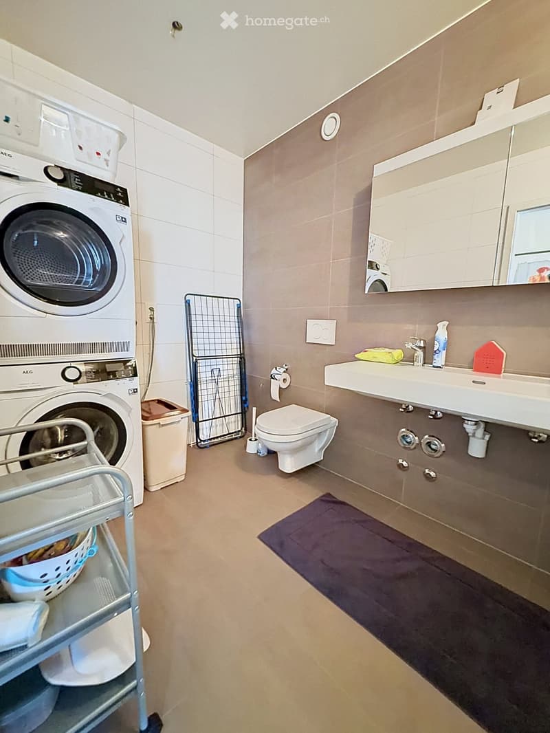 Modern bathroom with a toilet, sink, mirror, washing machine, dryer, laundry basket, and laundry rack.