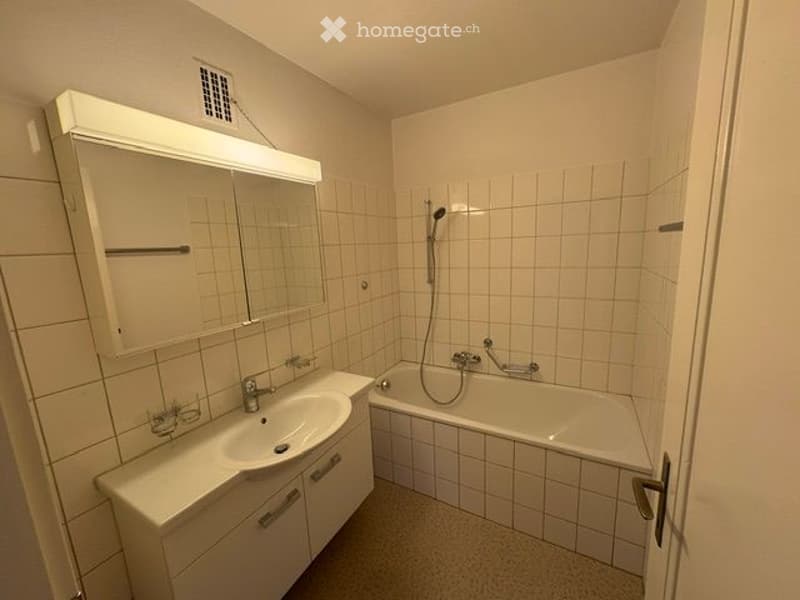 A small bathroom with white tiled walls and floor, featuring a sink with a cabinet, a bathtub, and a mirror with built-in lighting above it.