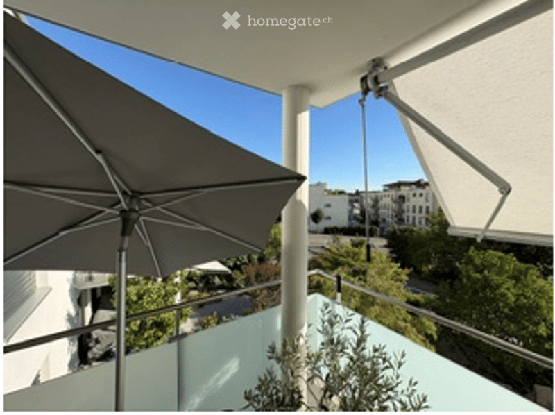 Balcony with a large umbrella, white railings, and a view of the city and greenery.