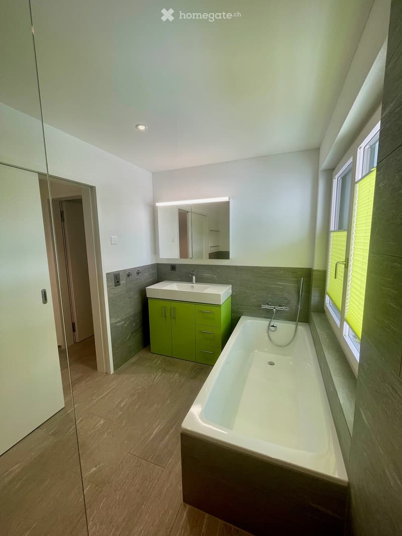 Bathroom. Modern bathroom with a bathtub, sink, green cabinet, and a large mirror above the sink. The room features light-colored wooden flooring and grey tiled walls.