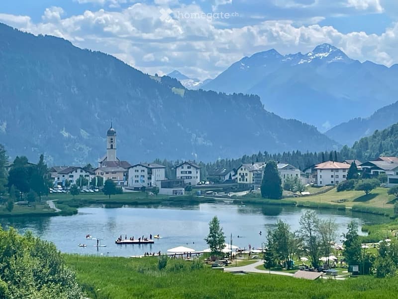 Scenic view of a village with a lake, surrounded by mountains, featuring houses, a church tower, and people enjoying the water and park areas.