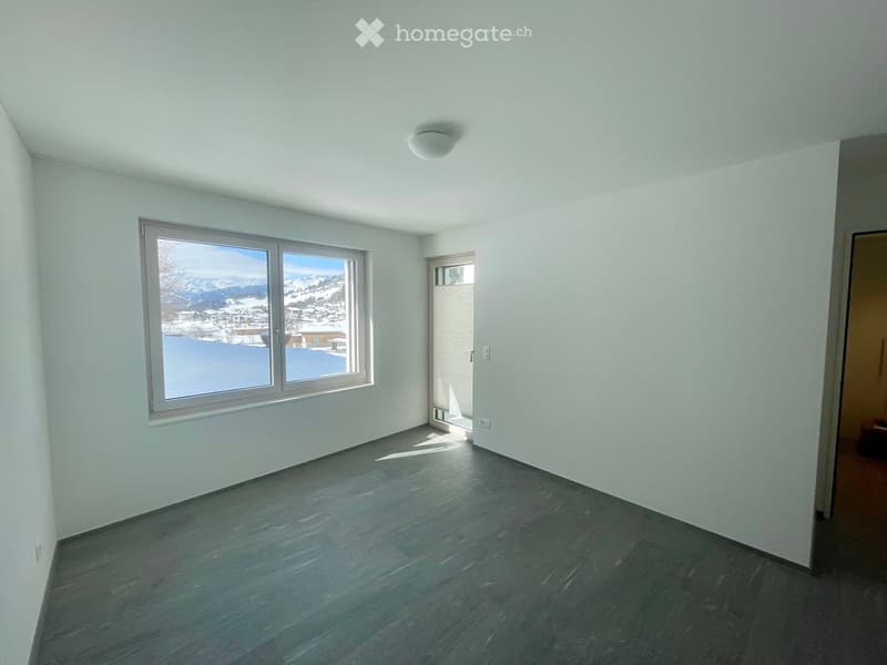 Unfurnished Room. Empty room with light-colored walls, dark wooden flooring, and large windows offering a view of snowy mountains and a town.
