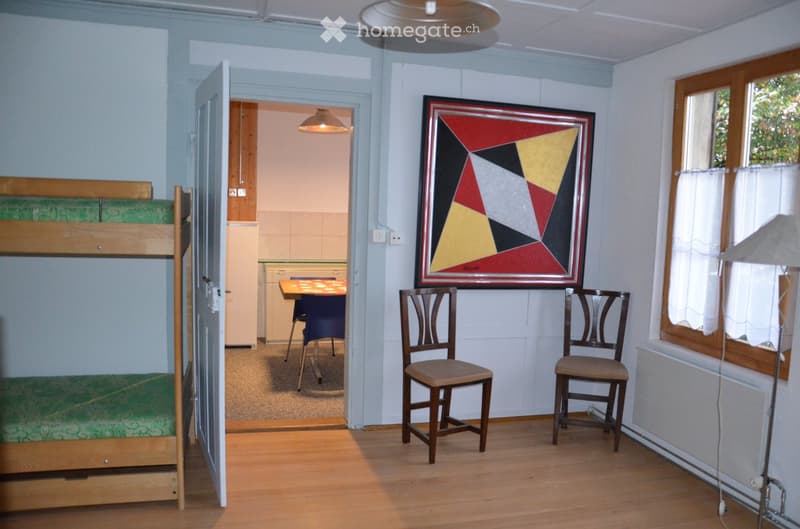 A furnished room with a bunk bed, two chairs, a small table, a picture frame on the wall, and a lamp near the window. The room has wooden flooring and light blue walls.