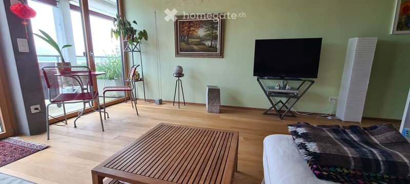 Bright living room with wooden flooring, large windows, a sofa, coffee table, chairs, a small table with a plant, a picture frame on the wall, and a speaker system.