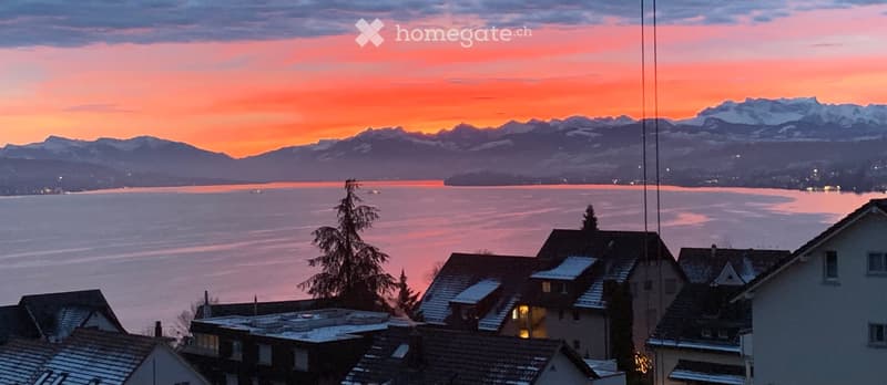 Panoramic view of a lake and snow-capped mountains at sunset, with rooftops of houses in the foreground.