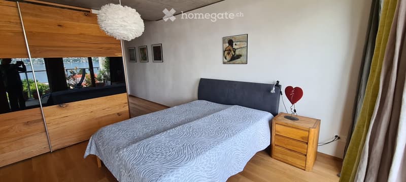 Cozy bedroom with a double bed, nightstand, lamp, and artwork on the wall. Large window offers a scenic view.