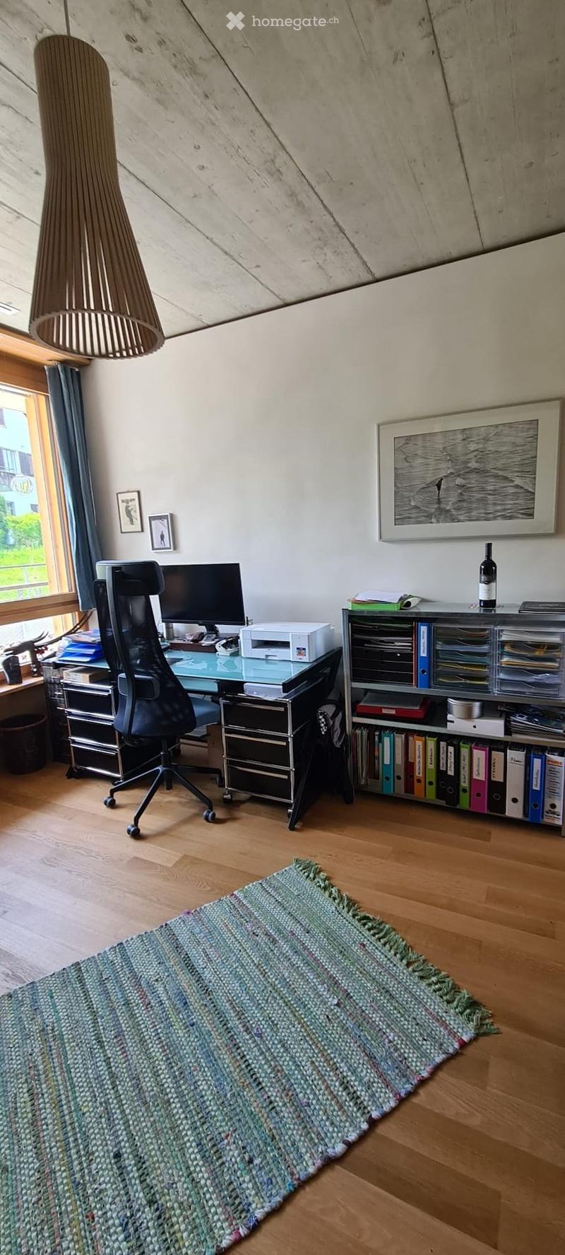 A modern, well-lit office space with a wooden floor, large window, and a woven rug. The room features a desk with a computer, printer, and office chair, along with a bookshelf filled with binders and books. A framed picture hangs on the wall, and a decora