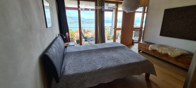 Modern bedroom with a large bed, nightstand, lamp, bench, and a scenic view through large windows and sliding doors leading to a balcony.