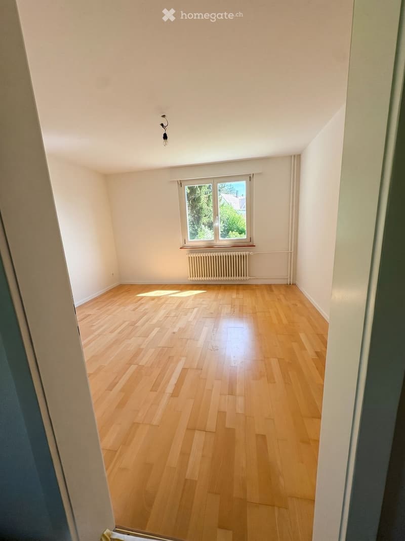 Empty room with wooden flooring, white walls, and a window with blinds. Natural light streams in, illuminating the space.