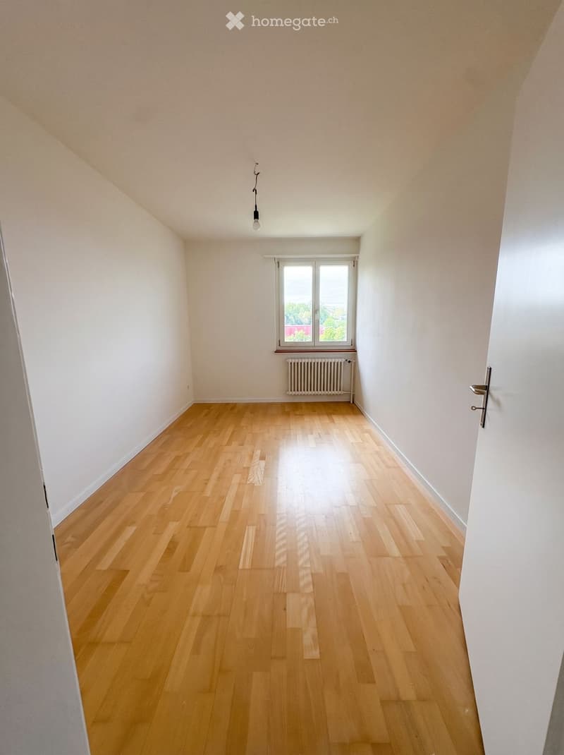 Empty room with light wooden flooring, white walls, a single window, and a radiator below it. The room has a simple, clean design with no furniture or decorations.
