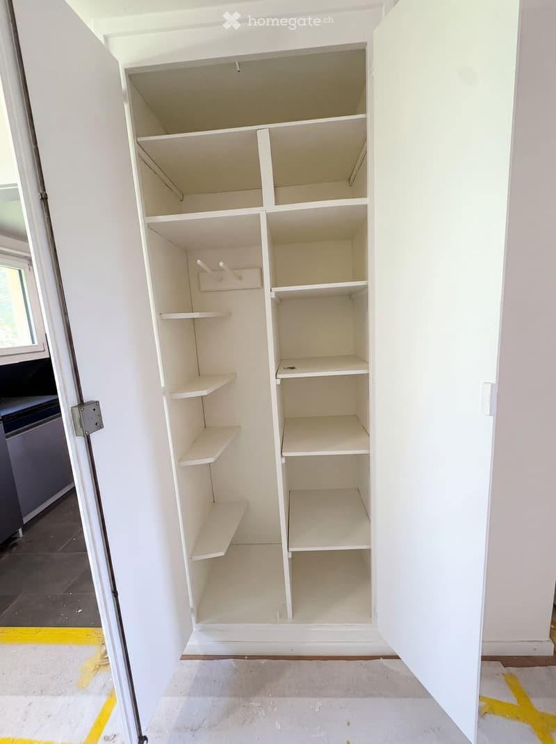 Empty closet with white shelves and a coat rack, located in a room with white walls and a tiled floor.