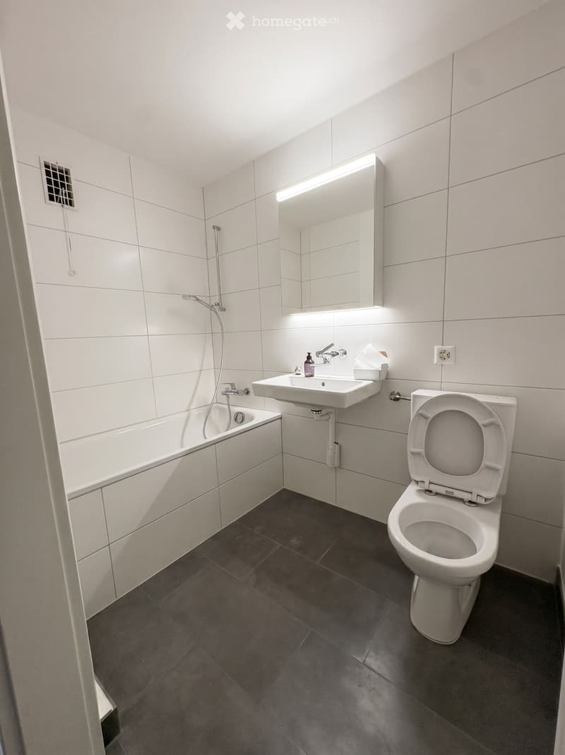 Modern bathroom with white tiles, a bathtub, a toilet, a sink, and a large mirror with built-in lighting.
