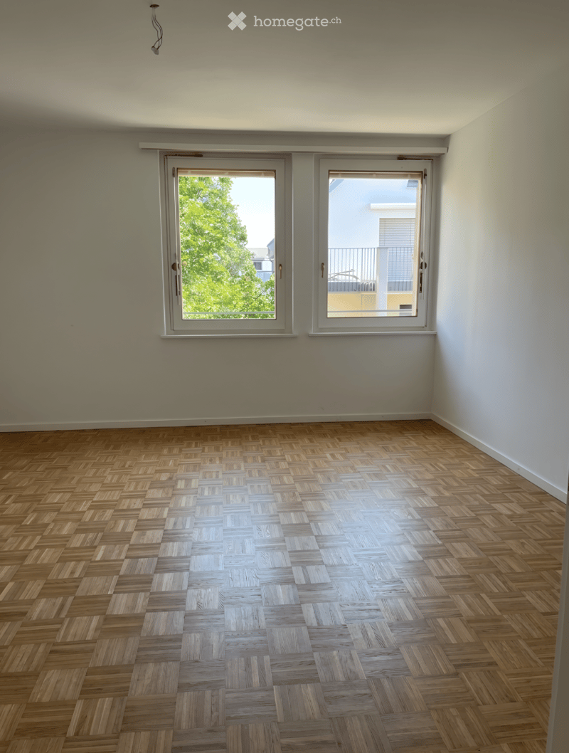 Empty room with wooden herringbone flooring and large windows offering a view of greenery and a neighboring building.