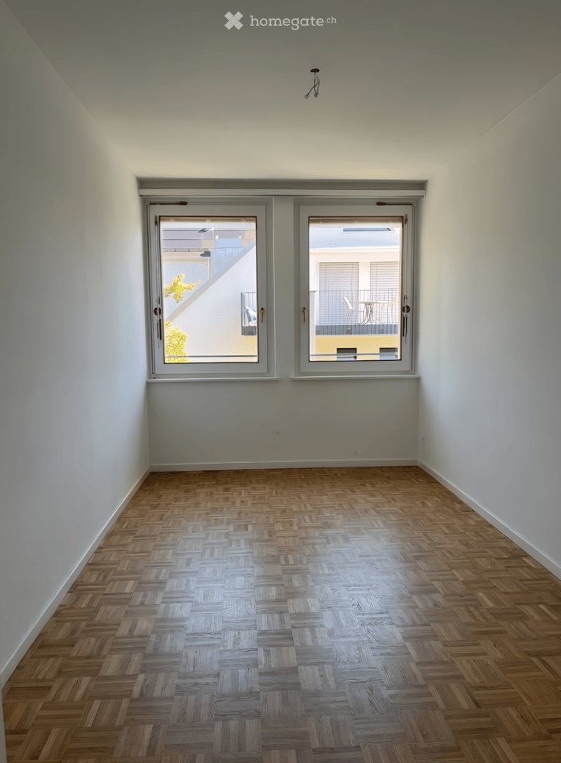 Empty room with wooden herringbone flooring, white walls, and large windows offering a view of a neighboring building and outdoor area.