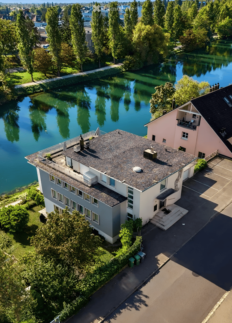 Aerial view of a modern two-story house with a grey roof and white walls, situated near a serene lake surrounded by lush greenery and trees.
