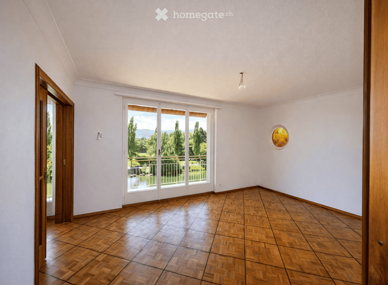 Empty room with wooden flooring, white walls, large sliding glass doors leading to a balcony with a scenic view, and a decorative round wall piece.