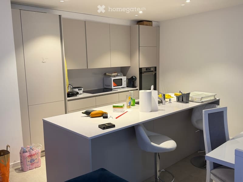 Modern kitchen with white cabinets, a central island, and various appliances including an oven and microwave. The island has a white countertop with a few items on it, and there are two stools at the island.
